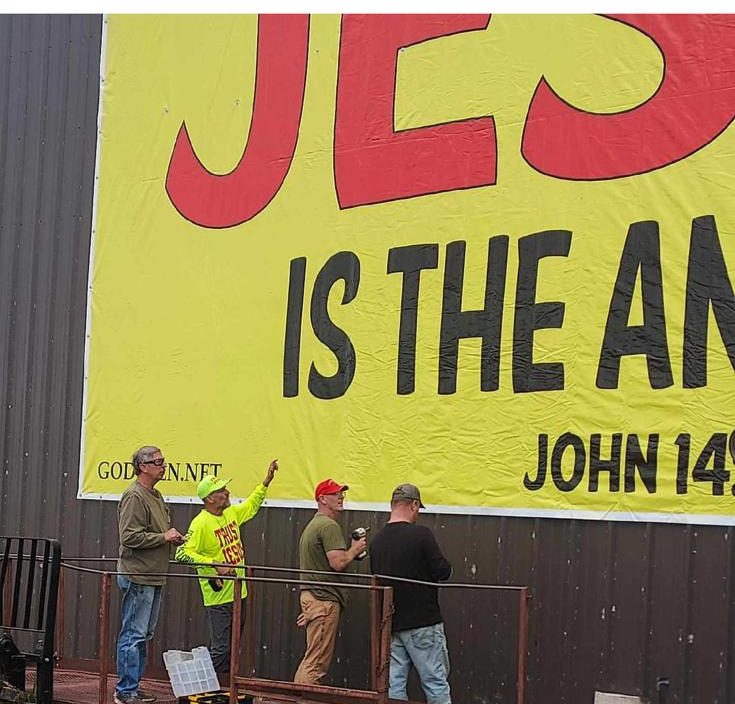 Four volunteers in TRUST JESUS shirts checking a newly hung JESUS IS THE ANSWER banner