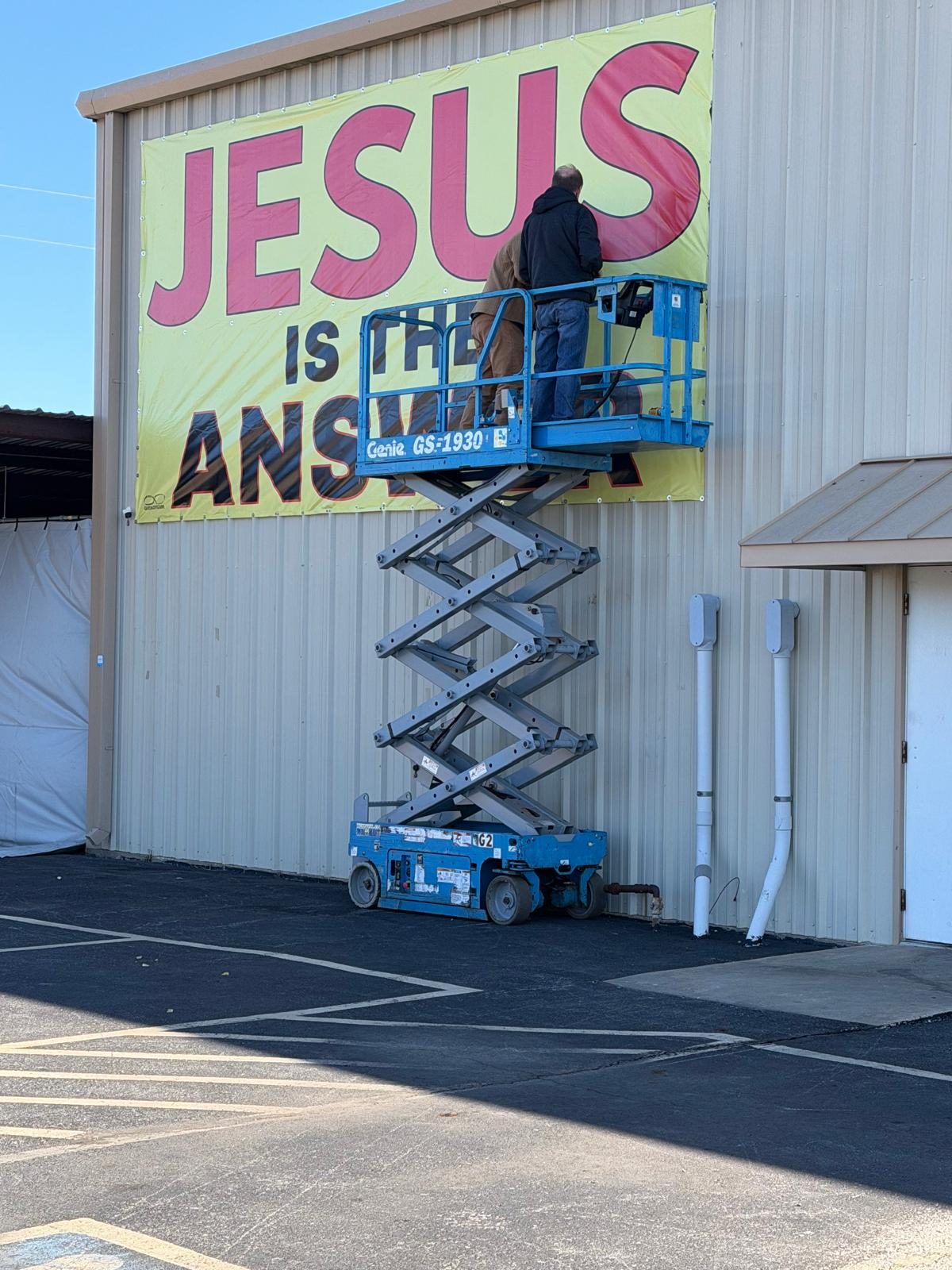 A man on a scissor lift smoothing out a JESUS IS THE ANSWER banner on a metal building